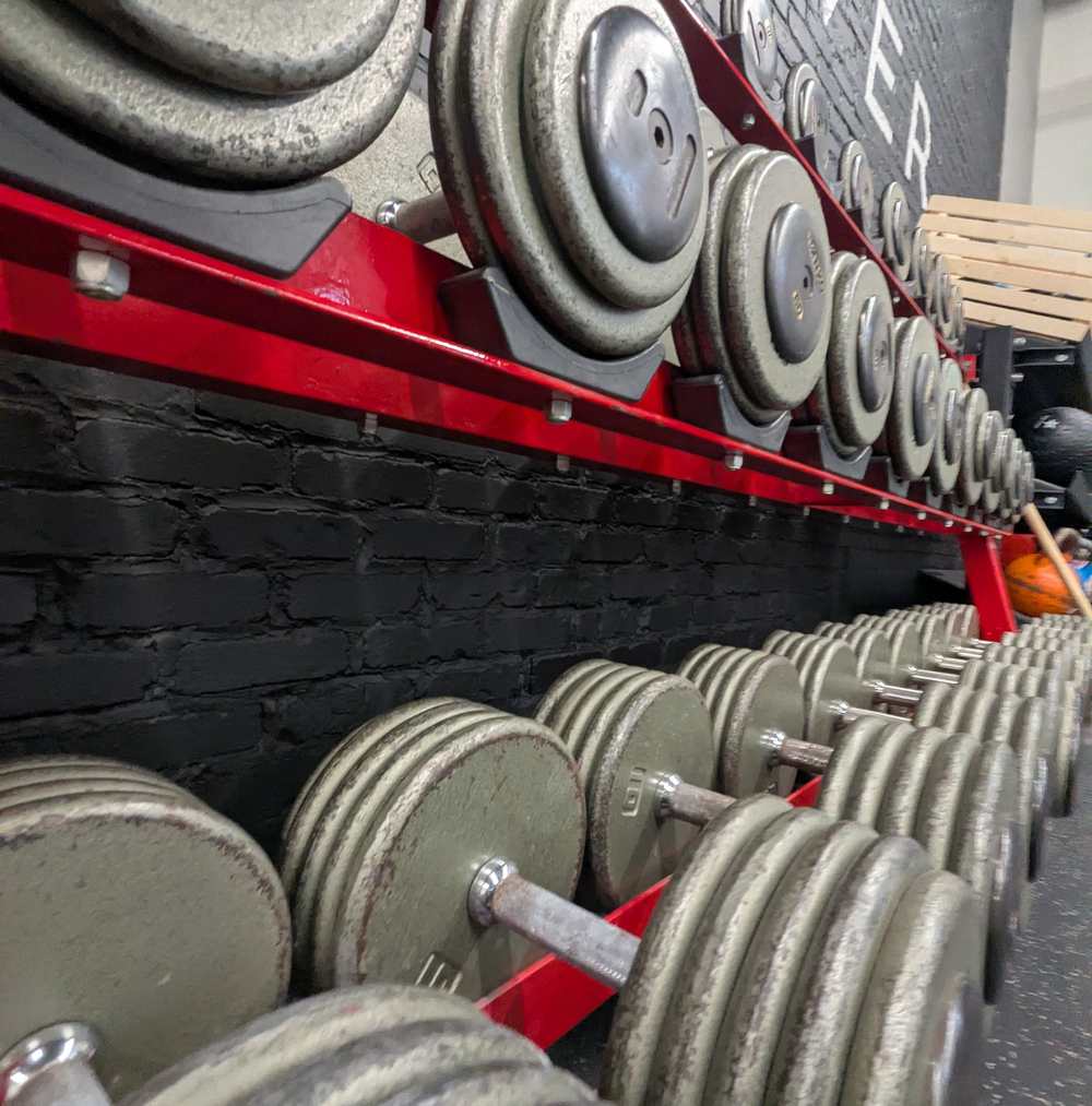 Rows of heavy dumbbells on red racks in front of the Better Forever logo wall